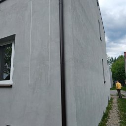 EKOENERGIA I AUTOMATYKA SPÓŁKA Z OGRANICZONĄ ODPOWIEDZIALNOŚCIĄ - Szara elewacja budynku po renowacji z brązową rynną, widoczne pionowe linie i okna. Osoba w tle na ścieżce.