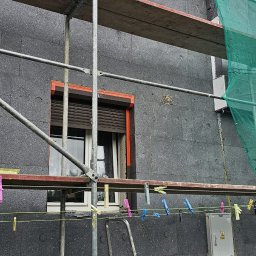 EKOENERGIA I AUTOMATYKA SPÓŁKA Z OGRANICZONĄ ODPOWIEDZIALNOŚCIĄ - Ocieplanie budynku styropianem grafitowym, widoczne rusztowanie i drabina. Okna z roletami w pomarańczowych obramowaniach. Na lince wiszą klamerki.