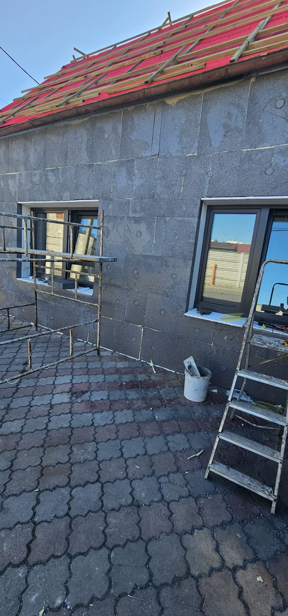 Ściana budynku w trakcie ocieplania styropianem grafitowym, widoczne okna, rusztowanie, drabina i wiadro z narzędziami. Czerwone elementy dachu.