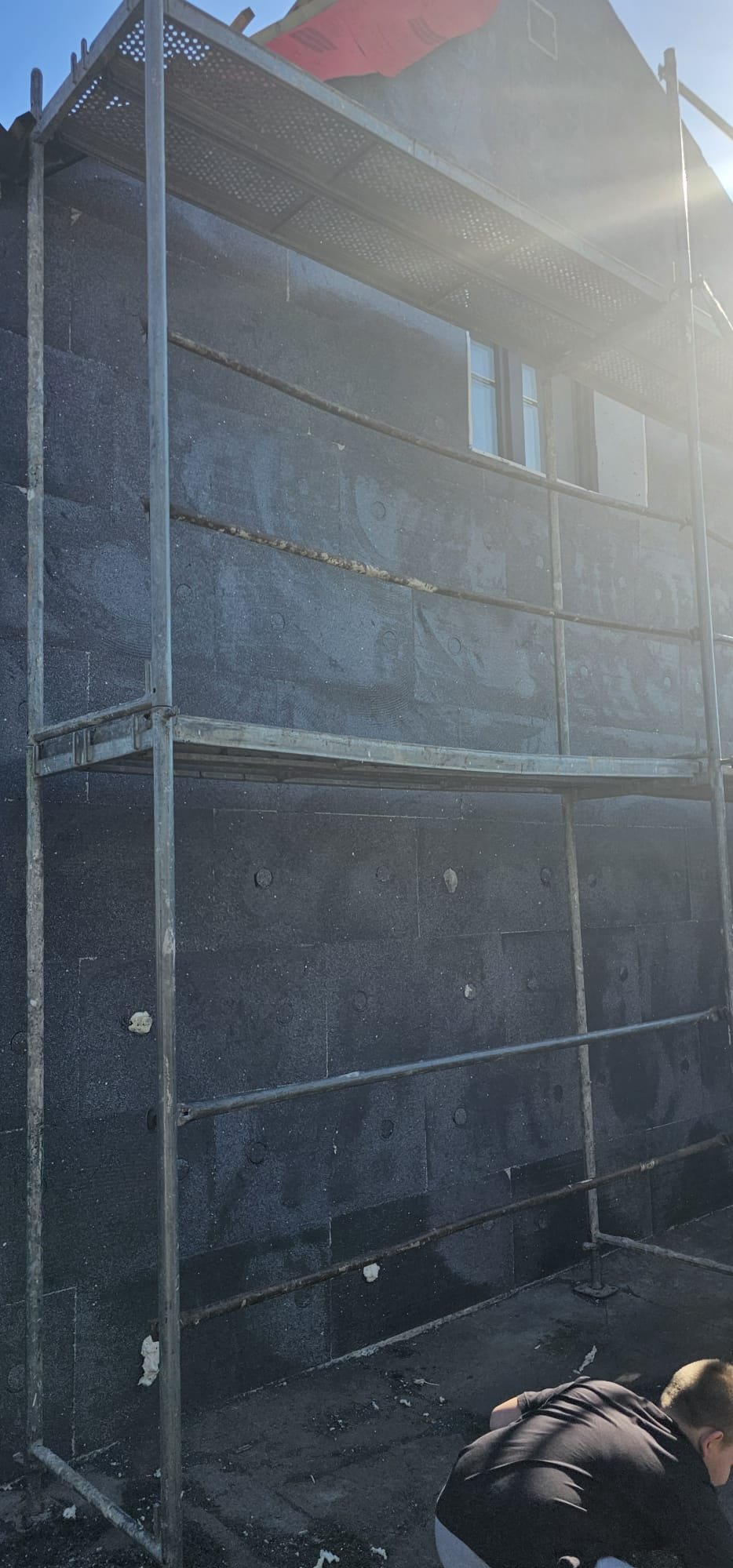 Elewacja domu w trakcie ocieplania czarnym styropianem, widoczne rusztowanie i fragment osoby w dolnym rogu. Etap prac z widocznymi mocowaniami.