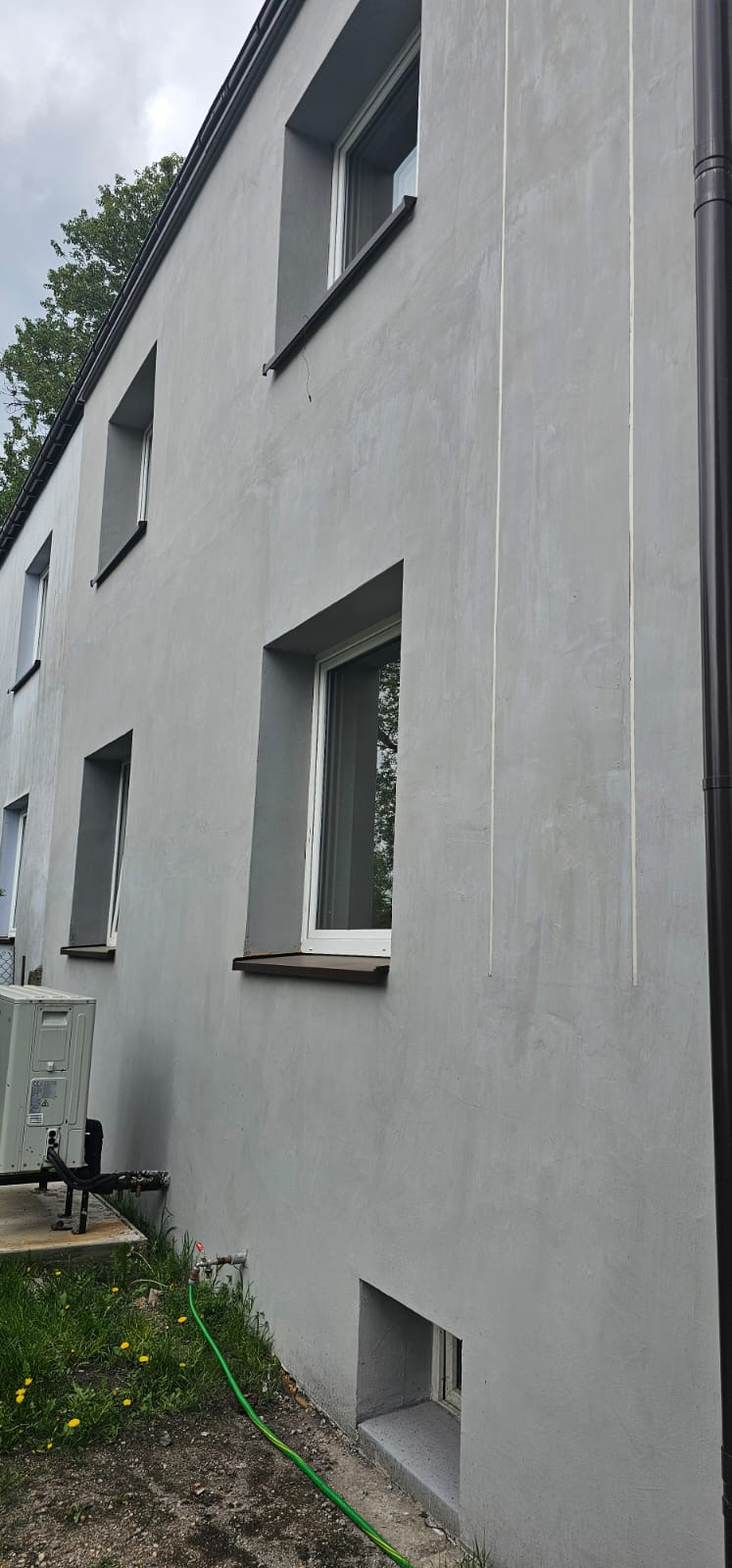 Szara elewacja budynku po renowacji z widocznymi pionowymi liniami, okna w szarych ramach, rura spustowa i klimatyzator na tle zieleni.