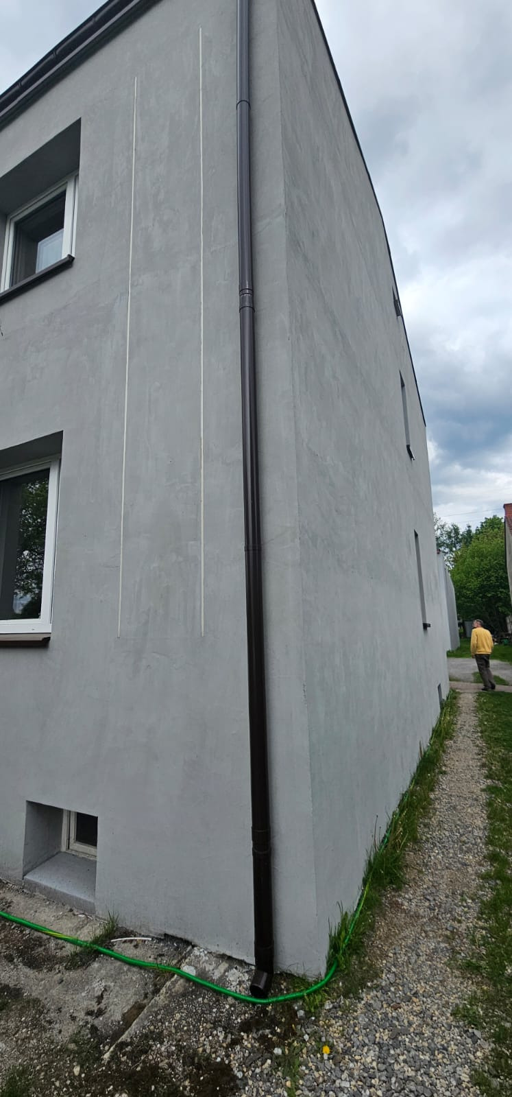 Szara elewacja budynku po renowacji z brązową rynną, widoczne pionowe linie i okna. Osoba w tle na ścieżce.