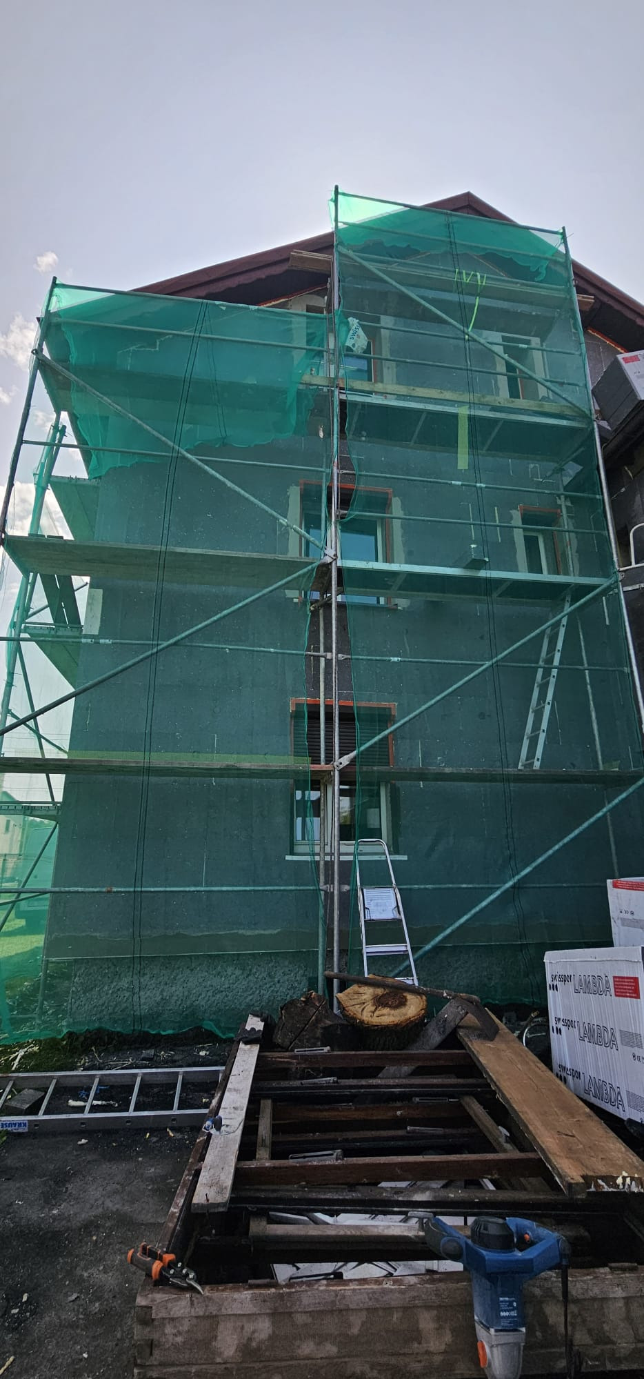 Dom w rusztowaniach, osłonięty zieloną siatką podczas prac elewacyjnych. Widoczne materiały izolacyjne i narzędzia na pierwszym planie.