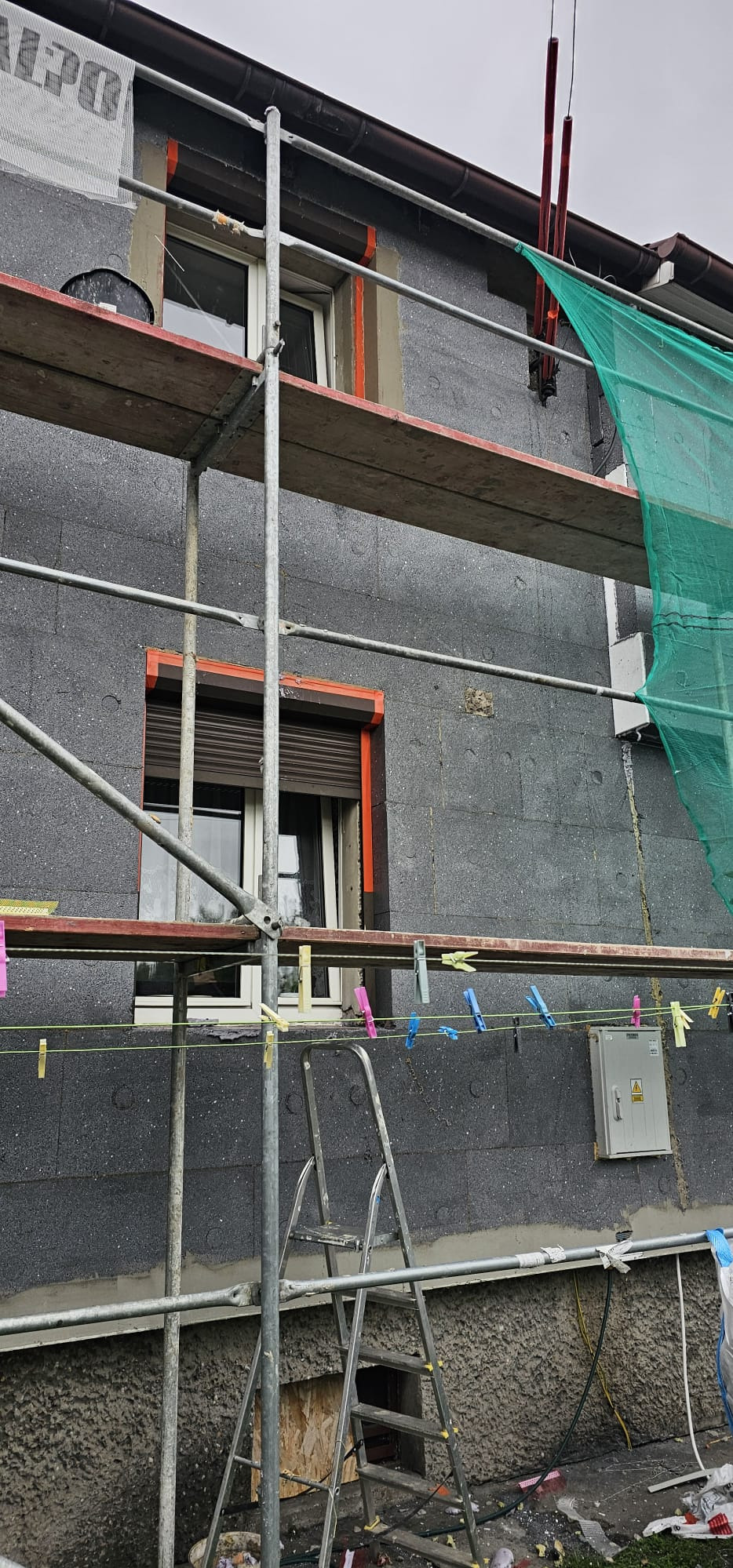 Ocieplanie budynku styropianem grafitowym, widoczne rusztowanie i drabina. Okna z roletami w pomarańczowych obramowaniach. Na lince wiszą klamerki.