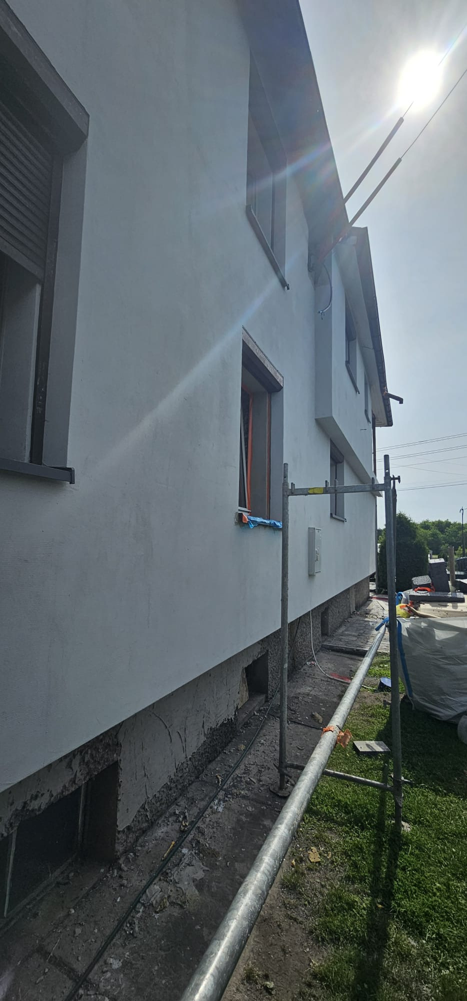 Boczny widok domu z ocieploną elewacją, widoczne rusztowania i niezakończone prace przy fundamentach, w tle błękitne niebo i rażące słońce.