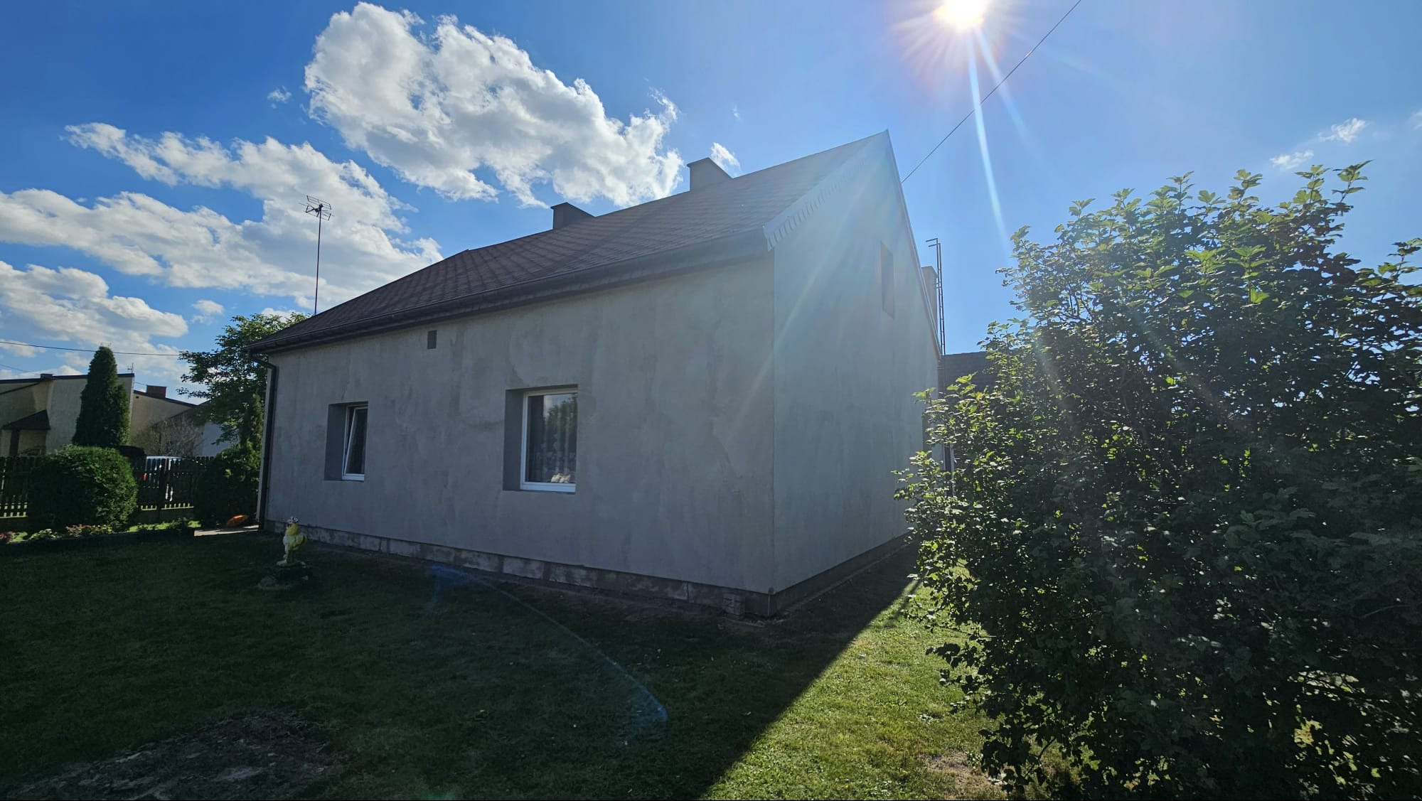 Dom jednorodzinny z szarą elewacją w słoneczny dzień. Widoczne dwa okna, brązowy dach i zielony trawnik. Na dachu antena.