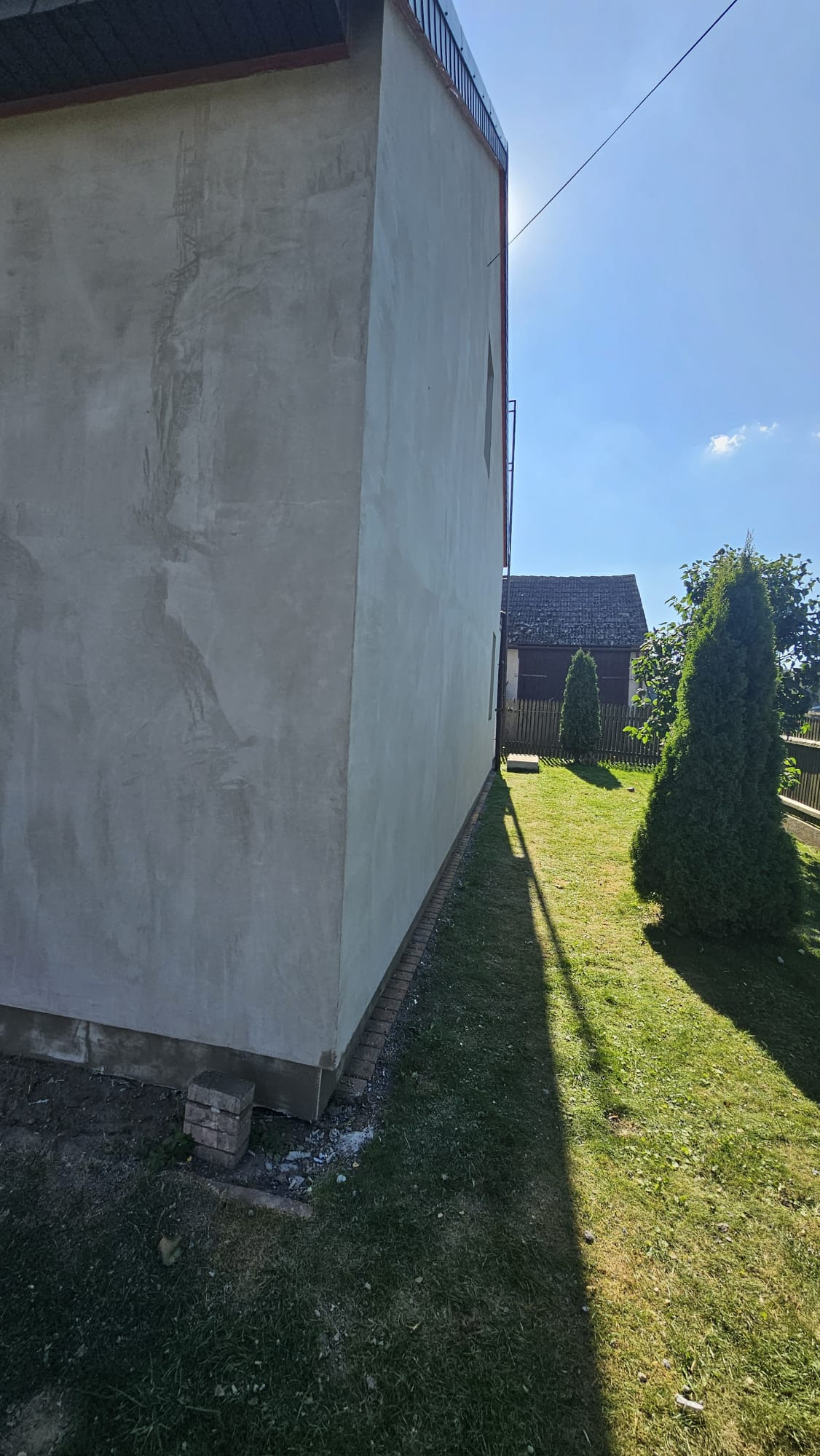 Ściana budynku po nałożeniu szarego tynku, widoczne niedoskonałości faktury, zieleń trawy i drzewa w tle. Słoneczny dzień.