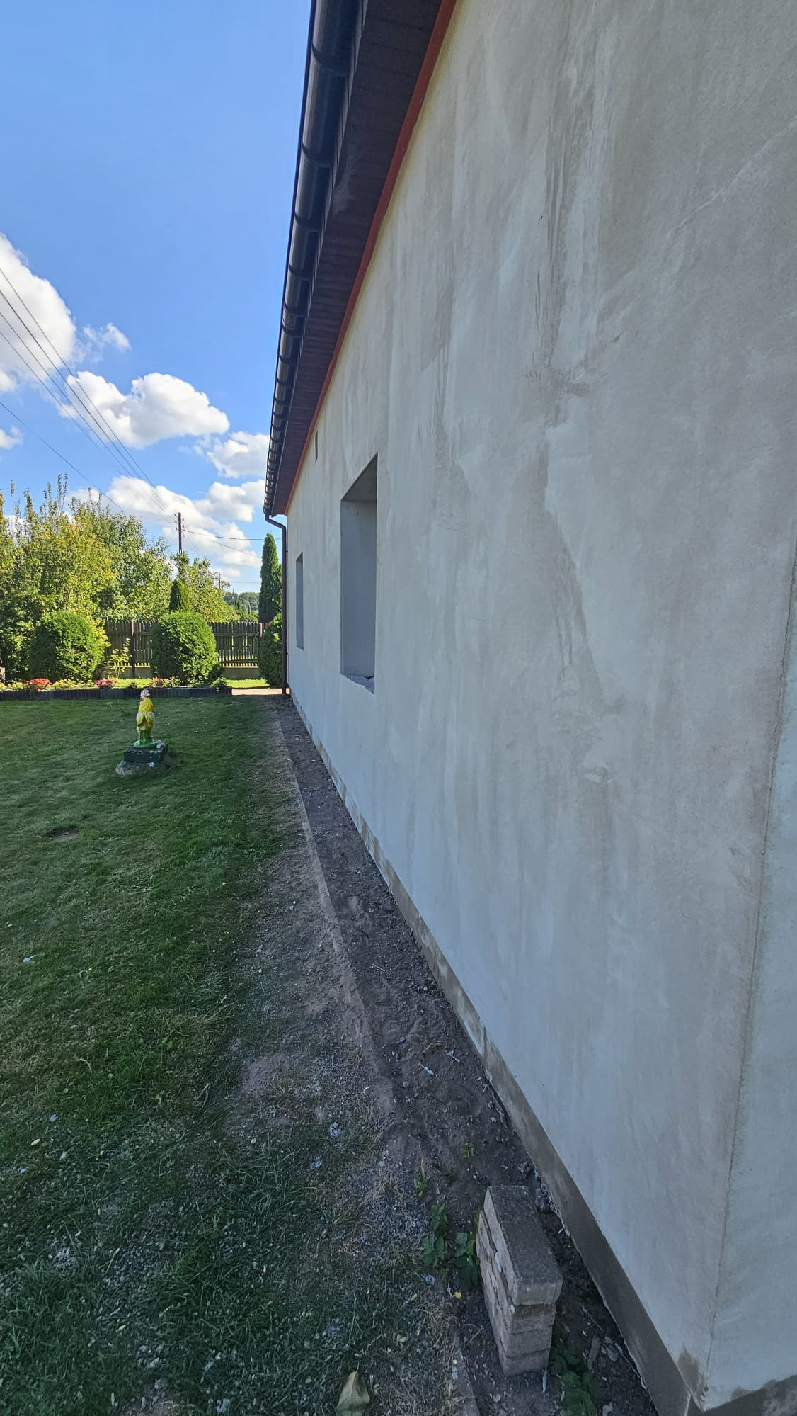 Ściana budynku po nałożeniu surowego tynku, widoczne okna bez ram i przygotowany teren wokół. Na trawniku stoi mała figurka ogrodowa.