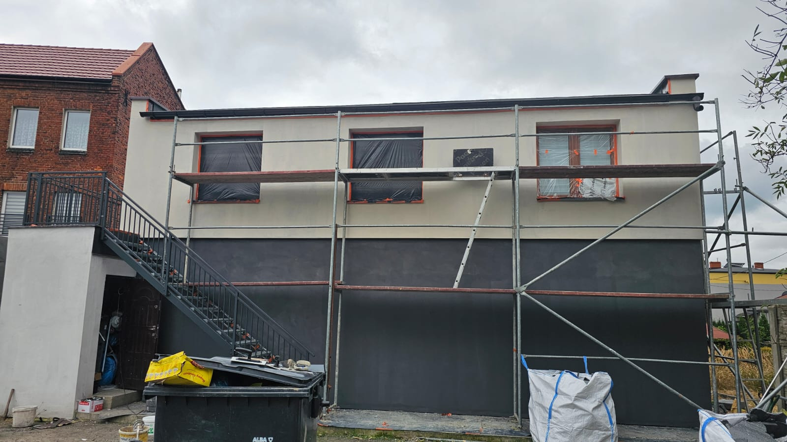 Dom w trakcie prac elewacyjnych: rusztowania, okna zabezpieczone folią, pomalowana dolna część fasady na ciemny kolor, górna w jasnym odcieniu. Widoczne narzędzia i materiały.