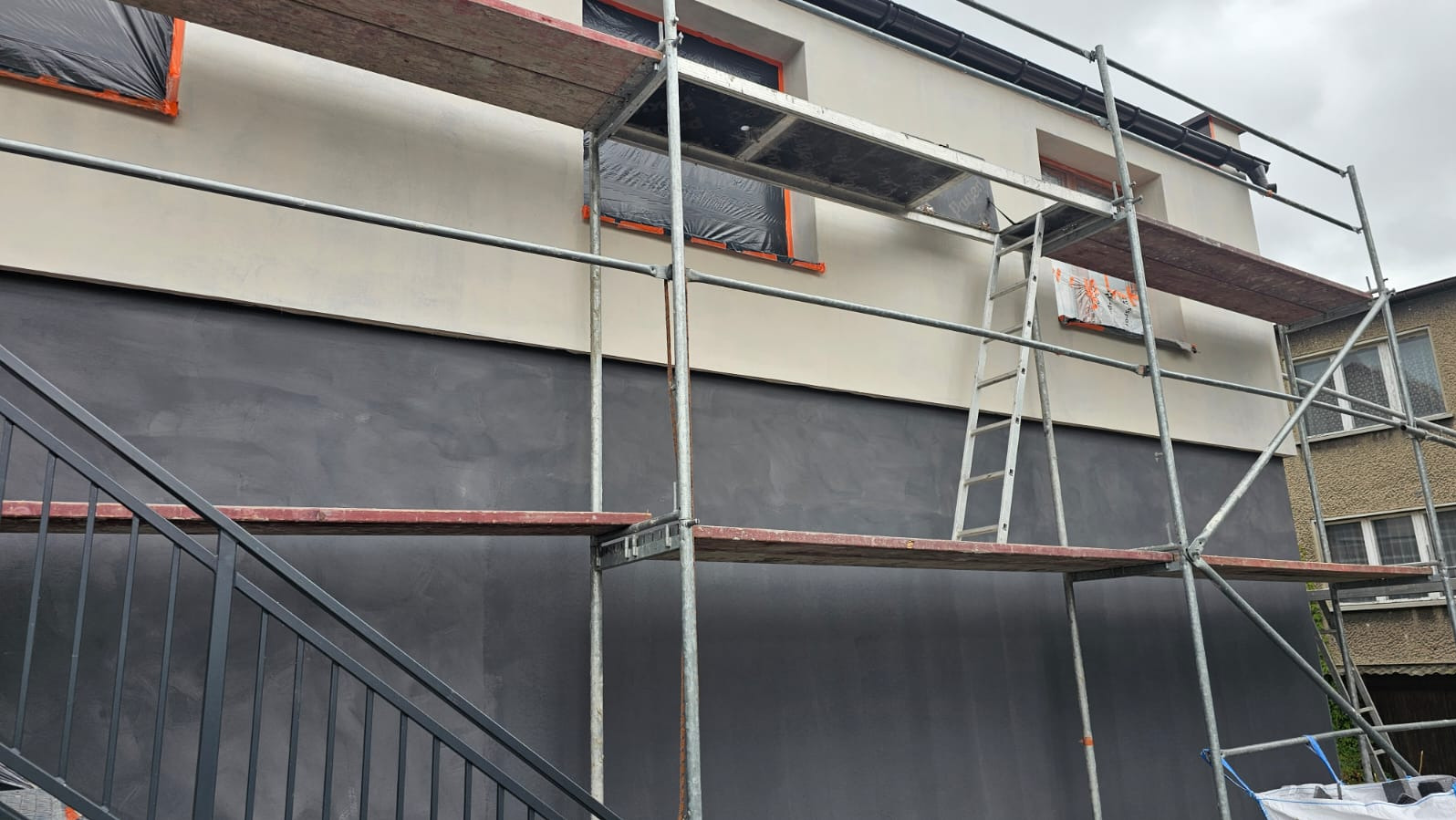 Dwukolorowa elewacja budynku w trakcie prac wykończeniowych. Widoczne rusztowania, okna zabezpieczone folią. Dolna część ściany pomalowana na szaro.