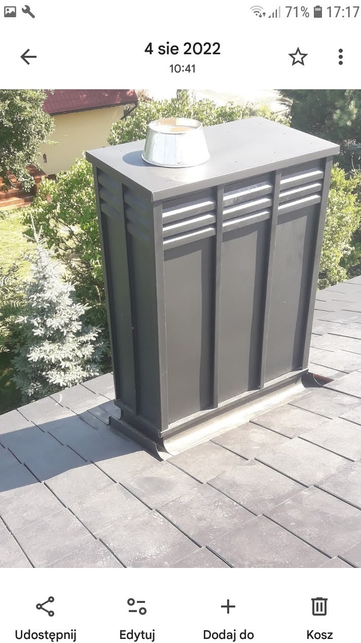 Szary komin wentylacyjny na dachu z szarej dachówki, z metalowym daszkiem. W tle zieleń drzew i fragment budynku. Ujęcie z góry, w słoneczny dzień.