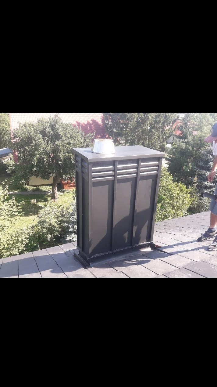 Szary komin wentylacyjny na dachu z dachówki bitumicznej, w tle zieleń drzew i fragment budynku. Widoczny fragment sylwetki osoby w prawym górnym rogu.