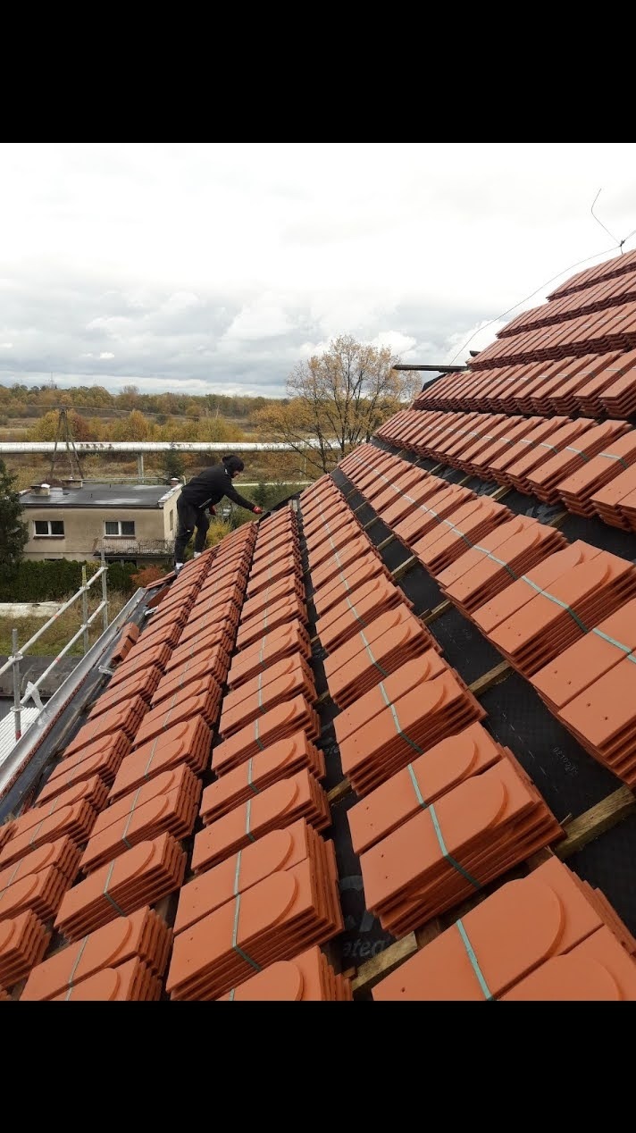 Układanie dachówki karpiówki na dachu przez dekarza. Widoczne przygotowane pakiety dachówek i fragment rusztowania. Tło: budynek i drzewa.