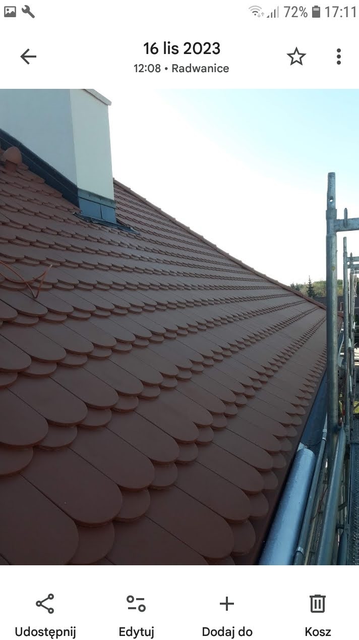 Brązowy dach kryty dachówką przypominającą łuskę, z widocznym kominem i fragmentem rusztowania. Ujęcie z dołu, w tle błękitne niebo.