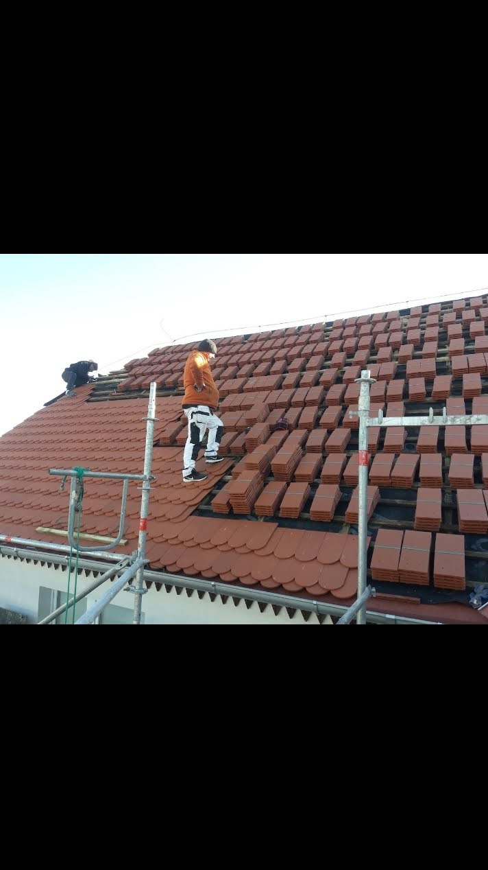 Układanie czerwonej dachówki karpiówki na dachu domu jednorodzinnego. Prace dekarskie prowadzone z rusztowania. Widoczni dekarze w trakcie pracy.