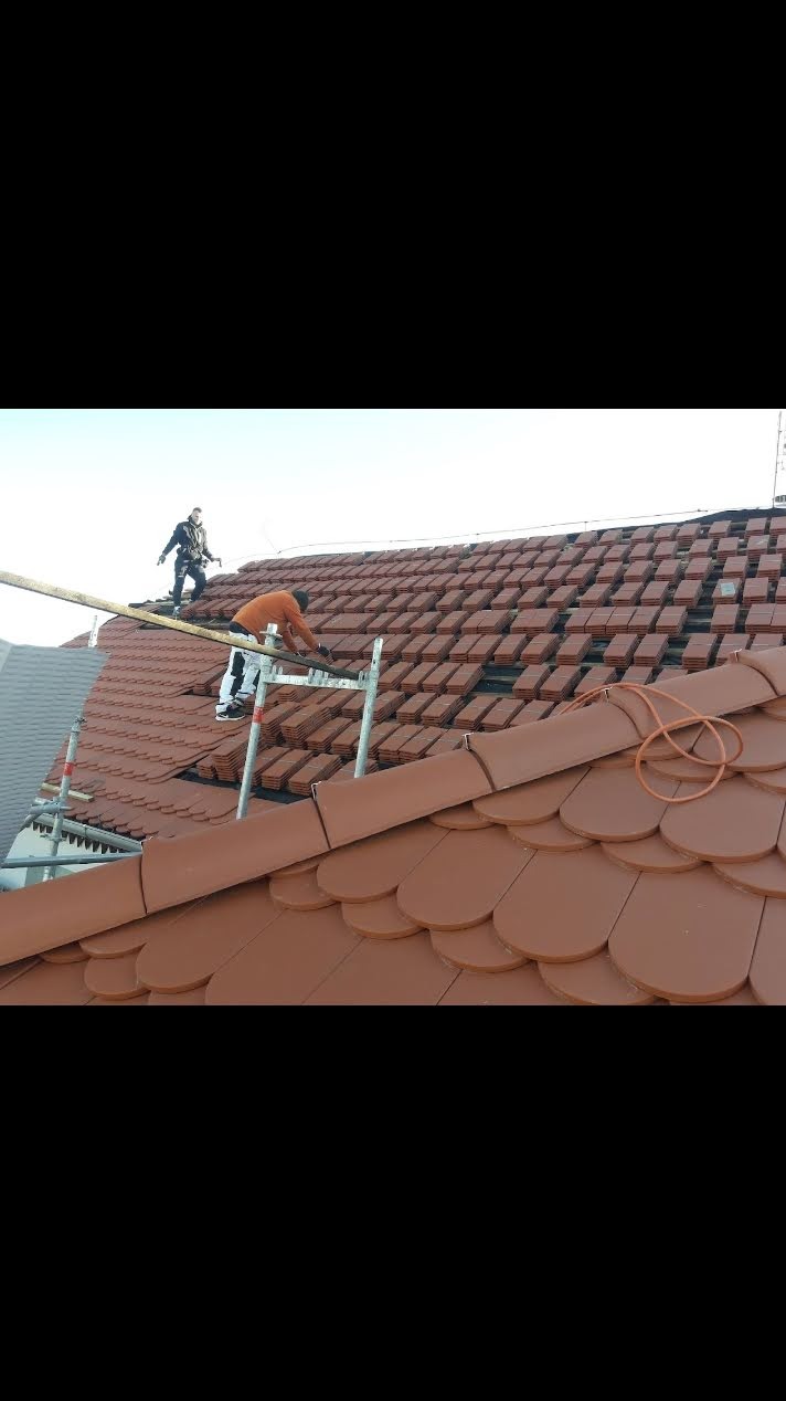 Układanie dachówki karpiówki na dachu w Siechnicach. Dwóch dekarzy na rusztowaniu. Częściowo pokryty dach, widoczne łaty i krokwie.