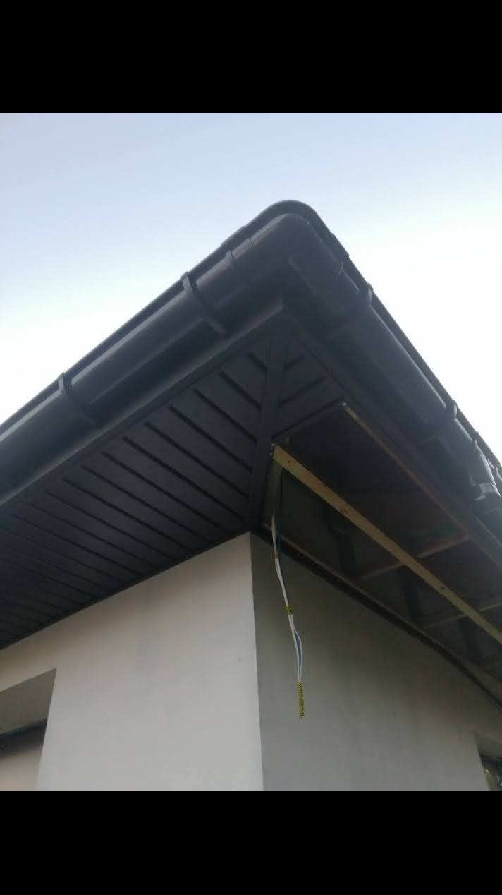 Ciemnoszara podbitka dachowa z rynnami na wykończonym budynku. Widoczne elementy konstrukcji dachu i wystające przewody.