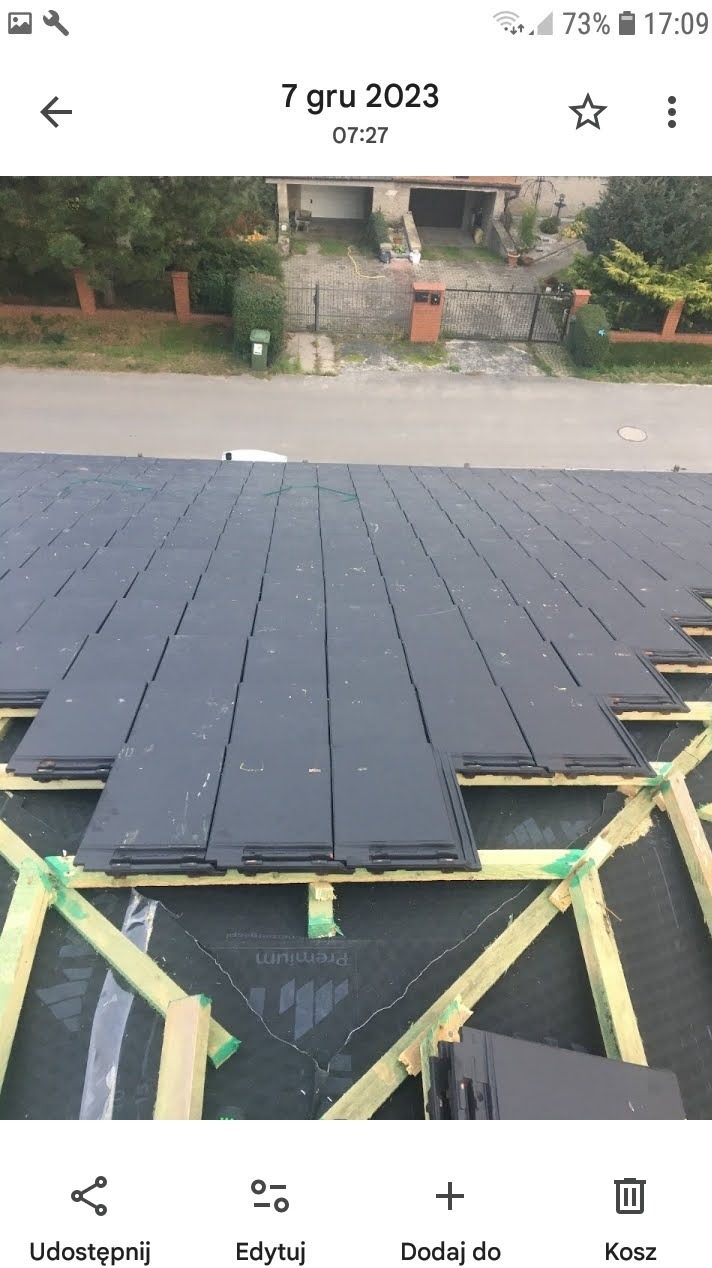 Układanie ciemnoszarej dachówki na drewnianej konstrukcji dachu. Widoczna membrana dachowa. W tle budynek mieszkalny i fragment ulicy.