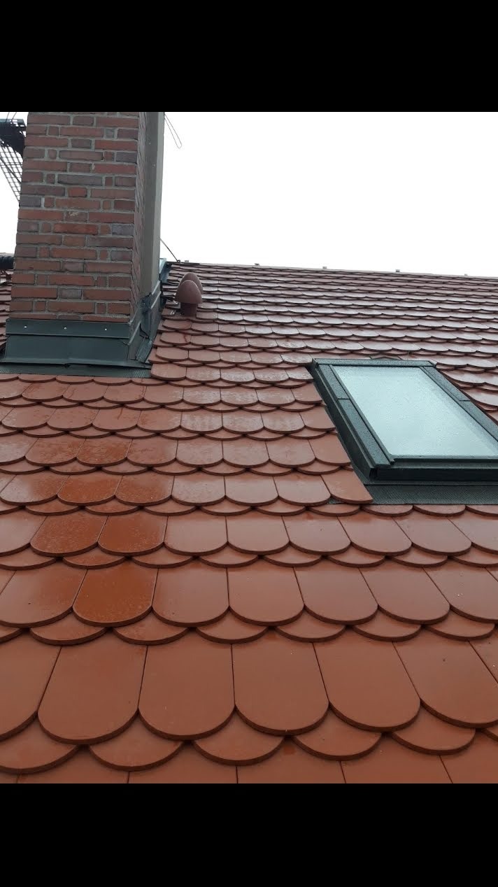 Dachówka karpiówka w odcieniach brązu, z widocznym oknem dachowym i kominem z czerwonej cegły. Lekko mokra powierzchnia sugeruje świeży opad deszczu.