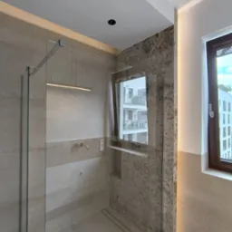 Nowoczesna kabina prysznicowa z lustrem w Siennicy. Beżowe płytki, szklana ściana, okno z widokiem na budynek. Minimalistyczny design, eleganckie wykończenie.