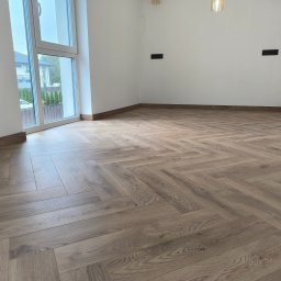 2B wykończenia wnętrz - Jasne, drewniane panele ułożone we wzór jodełki francuskiej w przestronnym, nowym pokoju z dużym oknem i białymi ścianami. Widoczne listwy przypodłogowe.