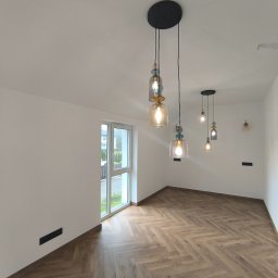 2B wykończenia wnętrz - Jasne wnętrze z parkietem w jodełkę, oświetlone przez designerskie lampy wiszące. Białe ściany i okno z widokiem na zieleń za oknem.