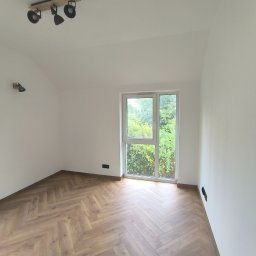 2B wykończenia wnętrz - Jasne, puste wnętrze z parkietem w jodełkę. Białe ściany, okno z widokiem na zieleń. Na suficie lampa z trzema reflektorami. Nowoczesne wykończenie.