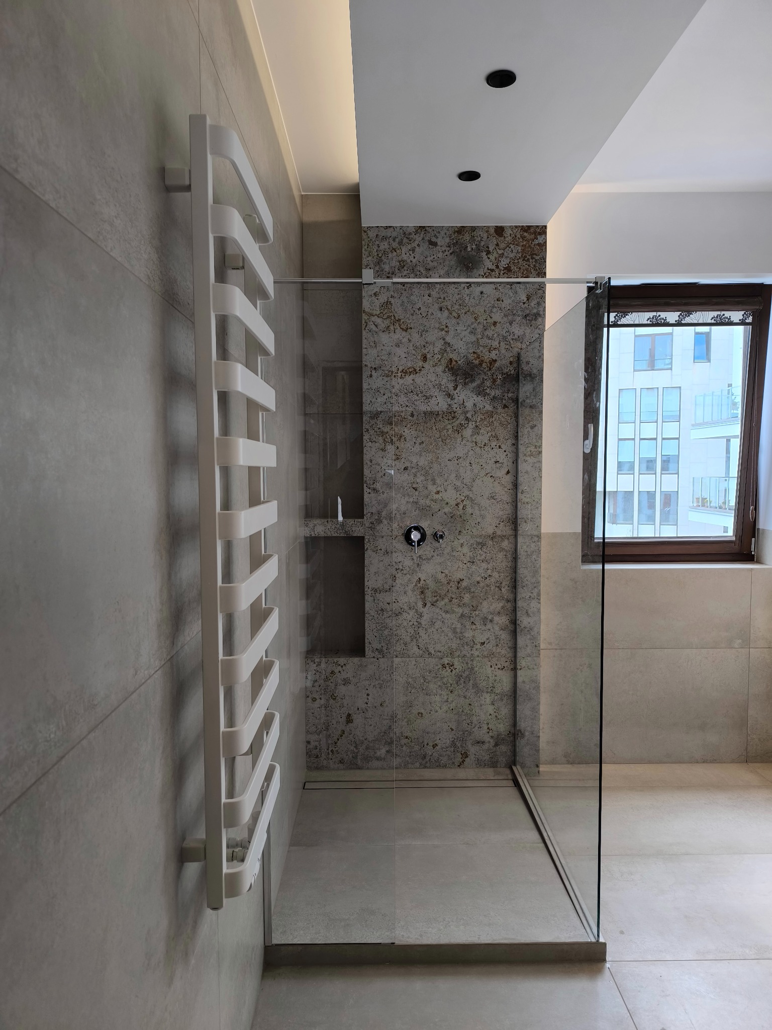 Nowoczesna łazienka z kabiną typu walk-in i grzejnikiem drabinkowym. Płytki w odcieniach szarości, okno z widokiem na budynek. Minimalistyczny design.