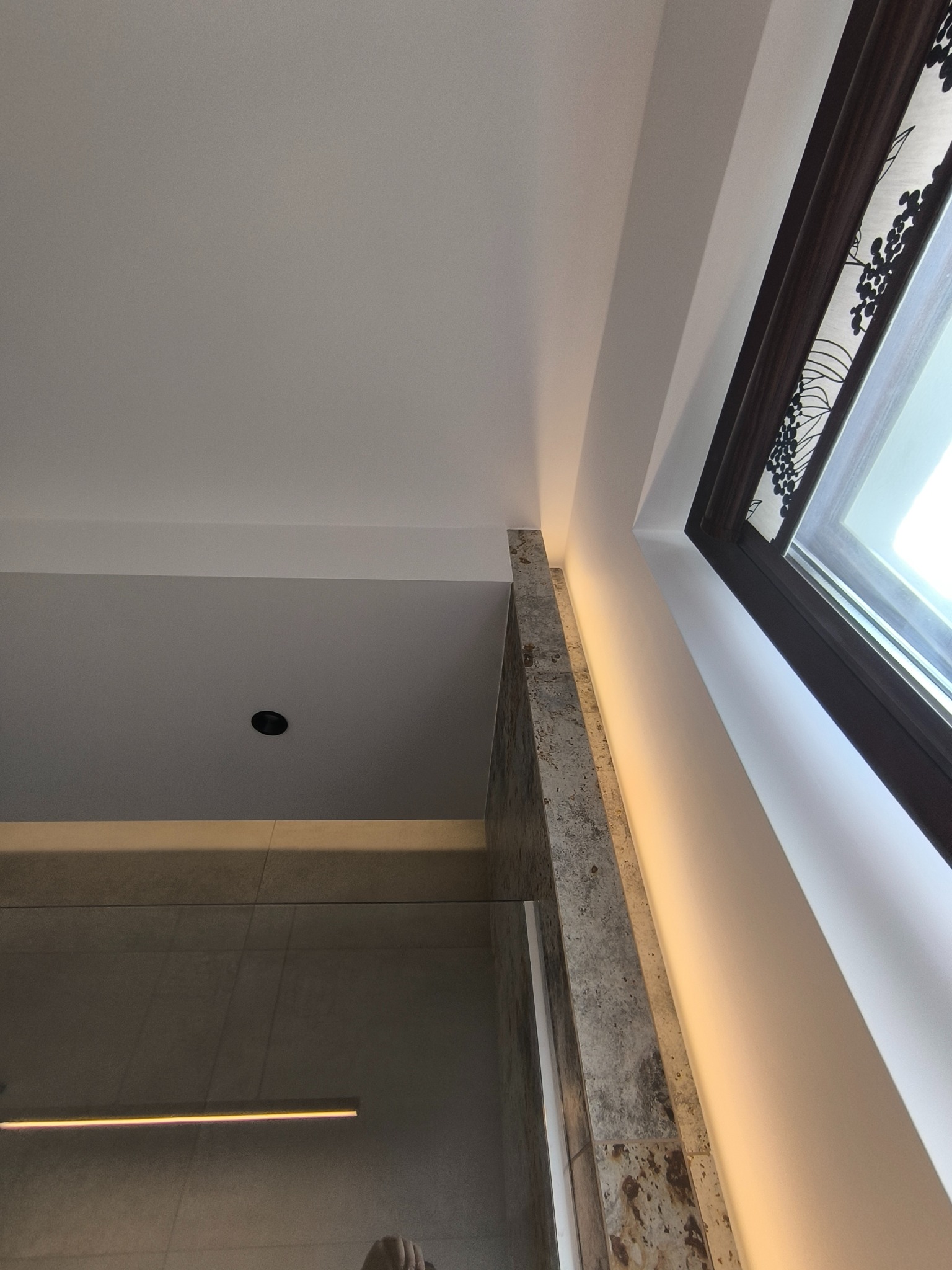 Nowoczesna łazienka z oknem dachowym i elementami z kamienia naturalnego. Widoczny fragment kabiny prysznicowej z szarymi płytkami i oświetleniem LED.