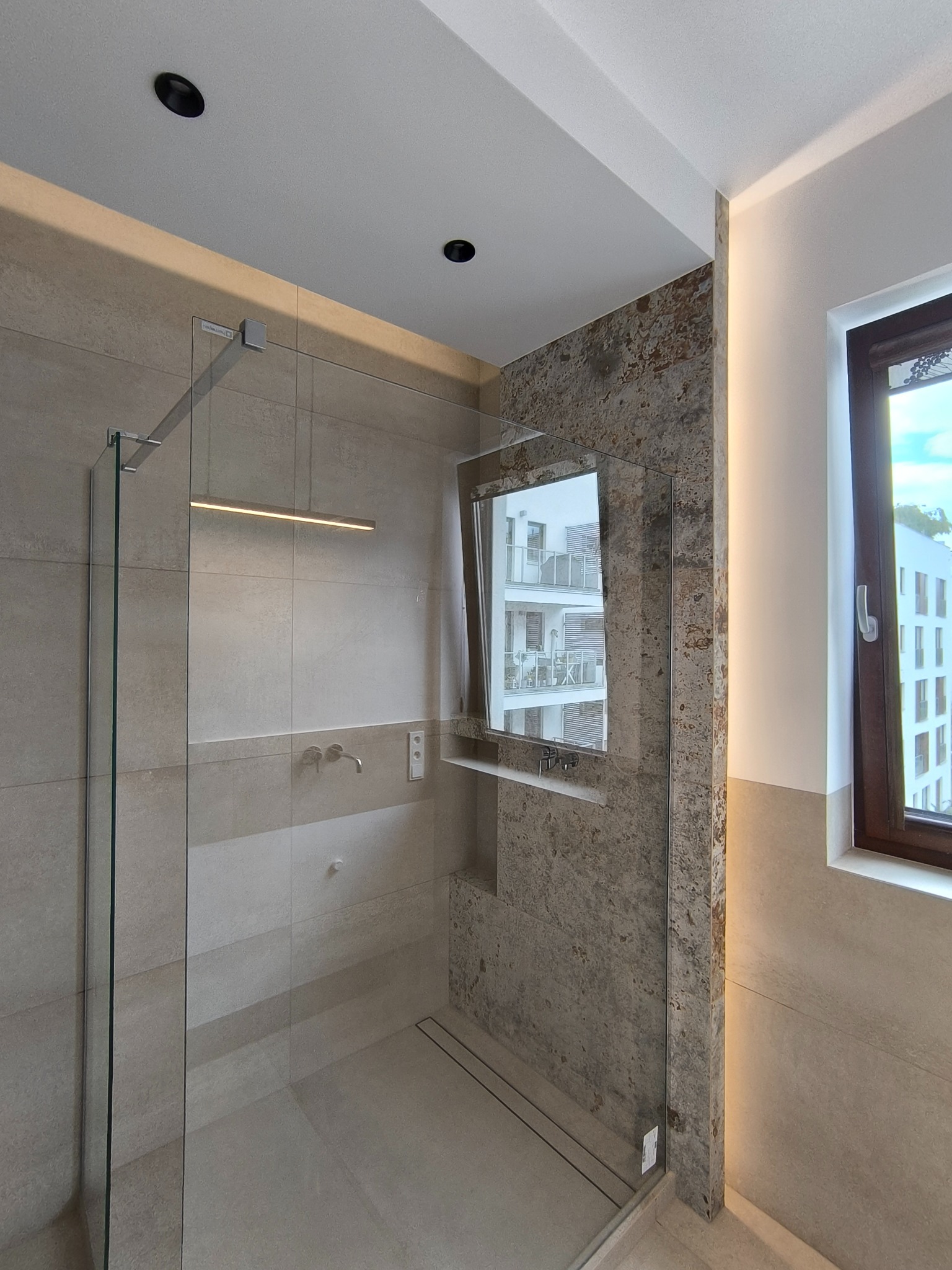 Nowoczesna kabina prysznicowa z lustrem w Siennicy. Beżowe płytki, szklana ściana, okno z widokiem na budynek. Minimalistyczny design, eleganckie wykończenie.