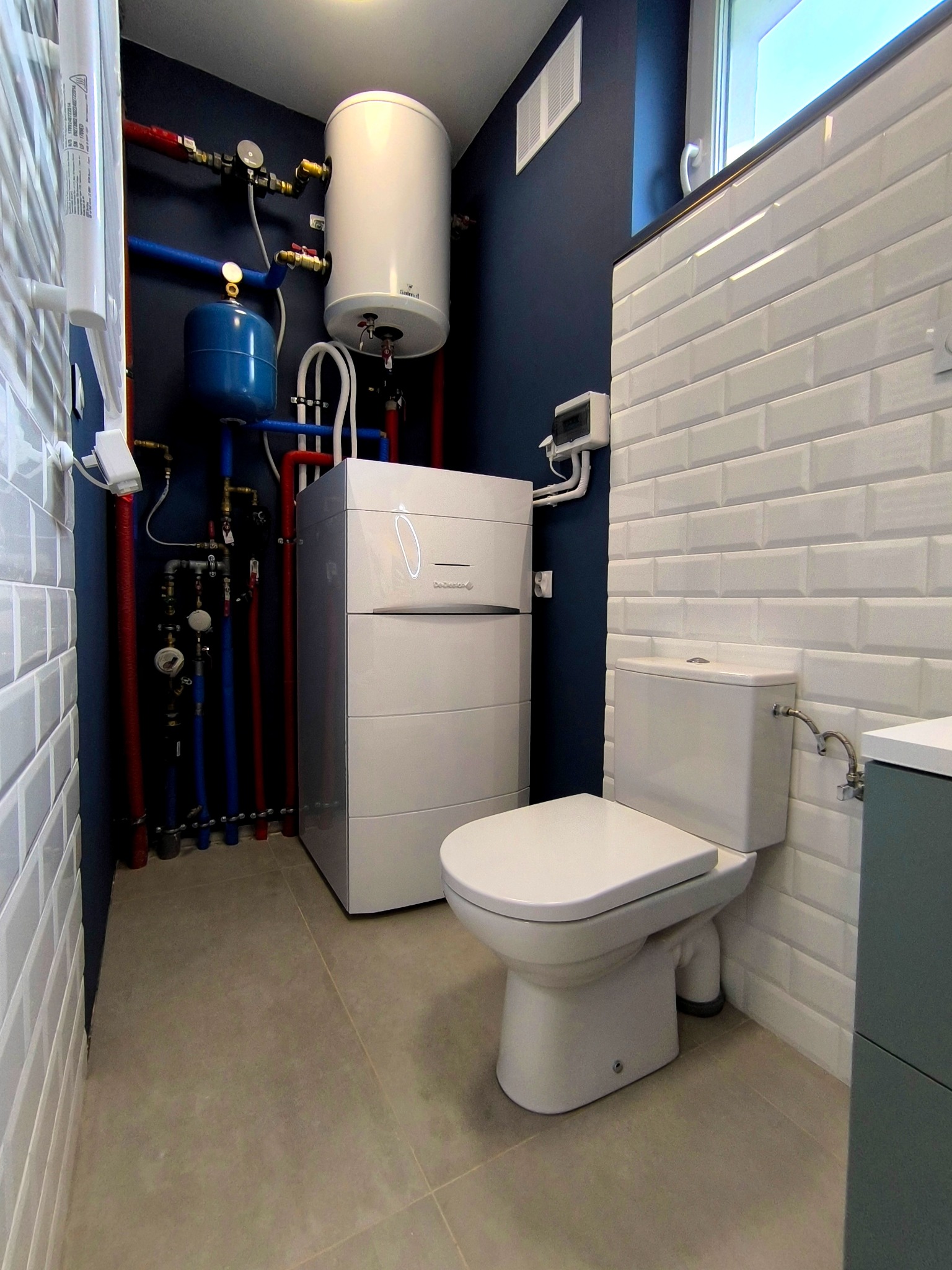 Nowoczesna łazienka z białymi, prostokątnymi płytkami na ścianach, szarą podłogą i nowym piecem grzewczym. Instalacja wodna z rurami w kolorach niebieskim i czerwonym.