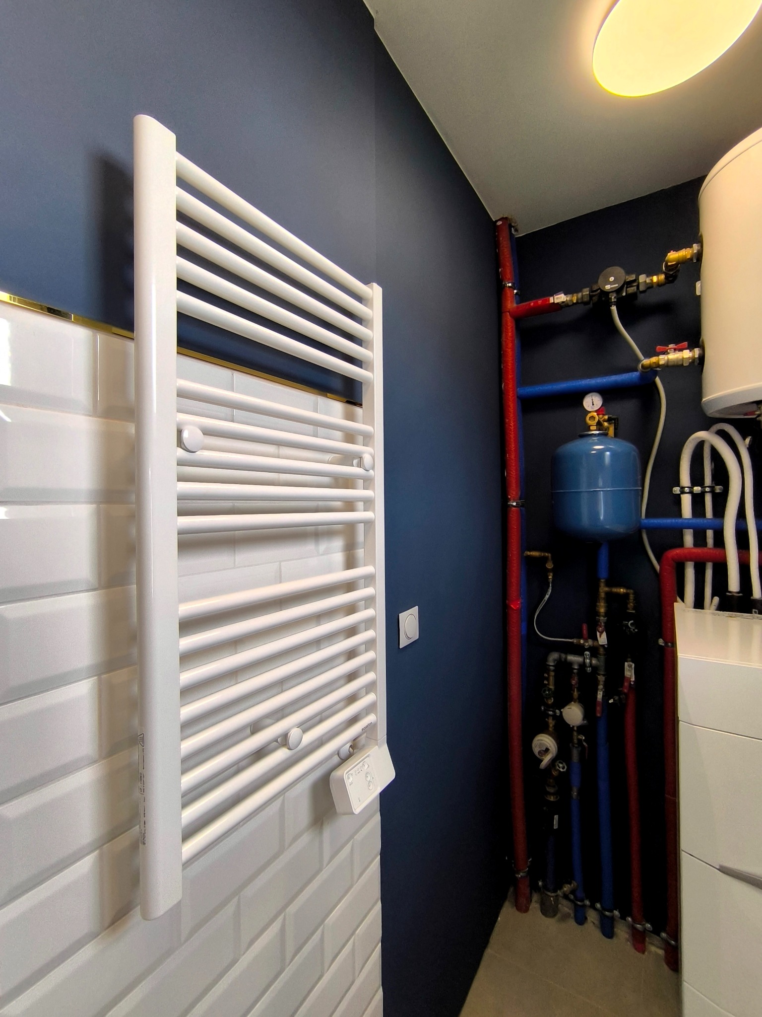 Nowoczesna łazienka z białym grzejnikiem drabinkowym na ścianie z białych płytek i instalacją grzewczą na tle granatowej ściany.