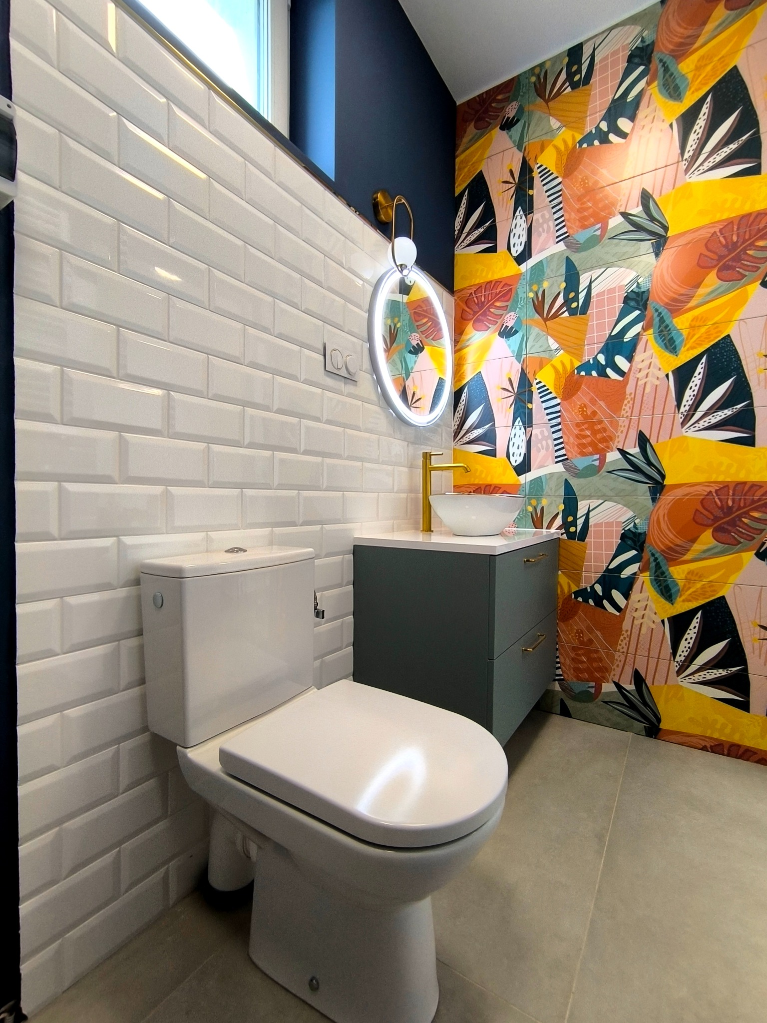Nowoczesna łazienka z białą armaturą, płytkami typu metro i akcentem kolorowej tapety w tropikalne wzory. Umywalka nablatowa i okrągłe lustro z podświetleniem LED.