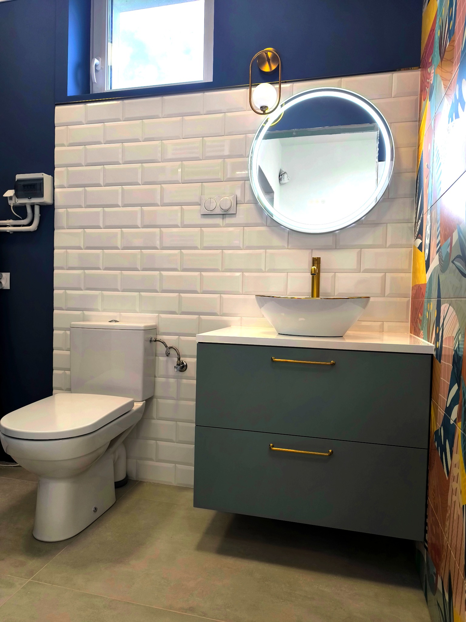 Nowoczesna łazienka w granatowo-białej tonacji z akcentami złota: okrągłe lustro z podświetleniem, umywalka nablatowa, białe płytki metro i kolorowe płytki dekoracyjne.