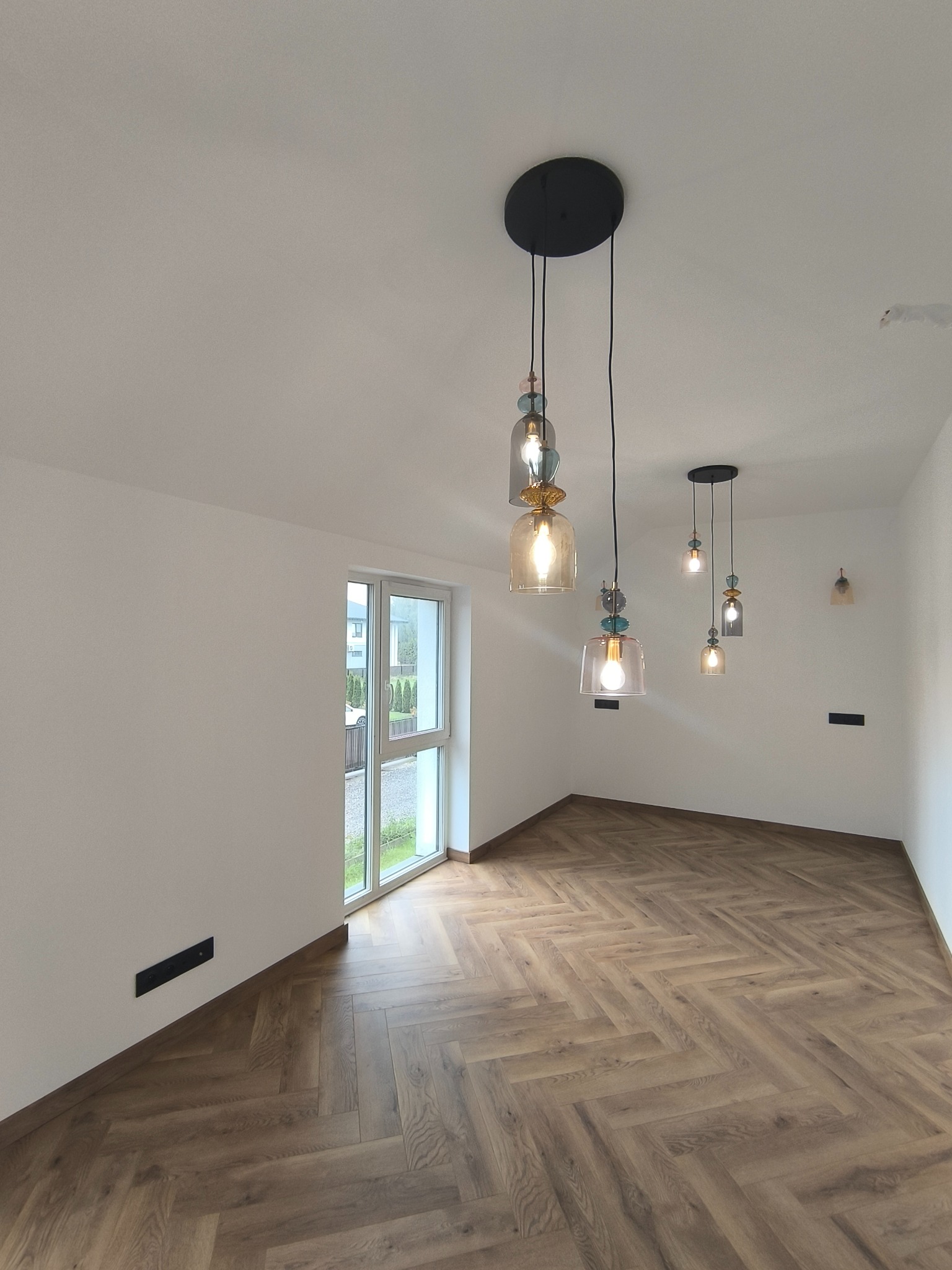 Jasne wnętrze z parkietem w jodełkę, oświetlone przez designerskie lampy wiszące. Białe ściany i okno z widokiem na zieleń za oknem.