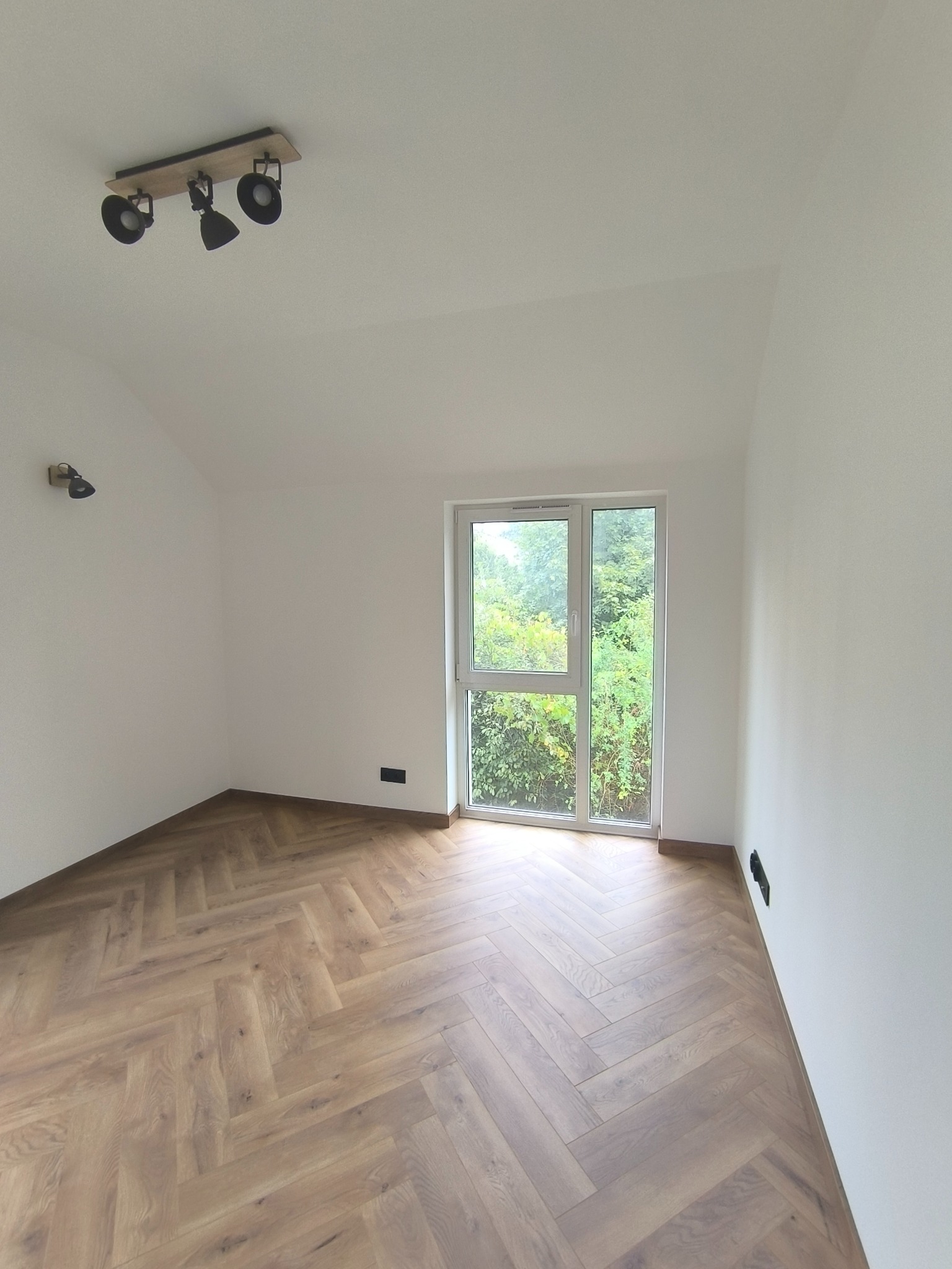 Jasne, puste wnętrze z parkietem w jodełkę. Białe ściany, okno z widokiem na zieleń. Na suficie lampa z trzema reflektorami. Nowoczesne wykończenie.