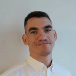 Portret młodego mężczyzny w białej koszuli na tle jasnej ściany. Krótkie, ciemne włosy, lekki uśmiech, wzrok skierowany w obiektyw. Dobre oświetlenie, naturalny wygląd.