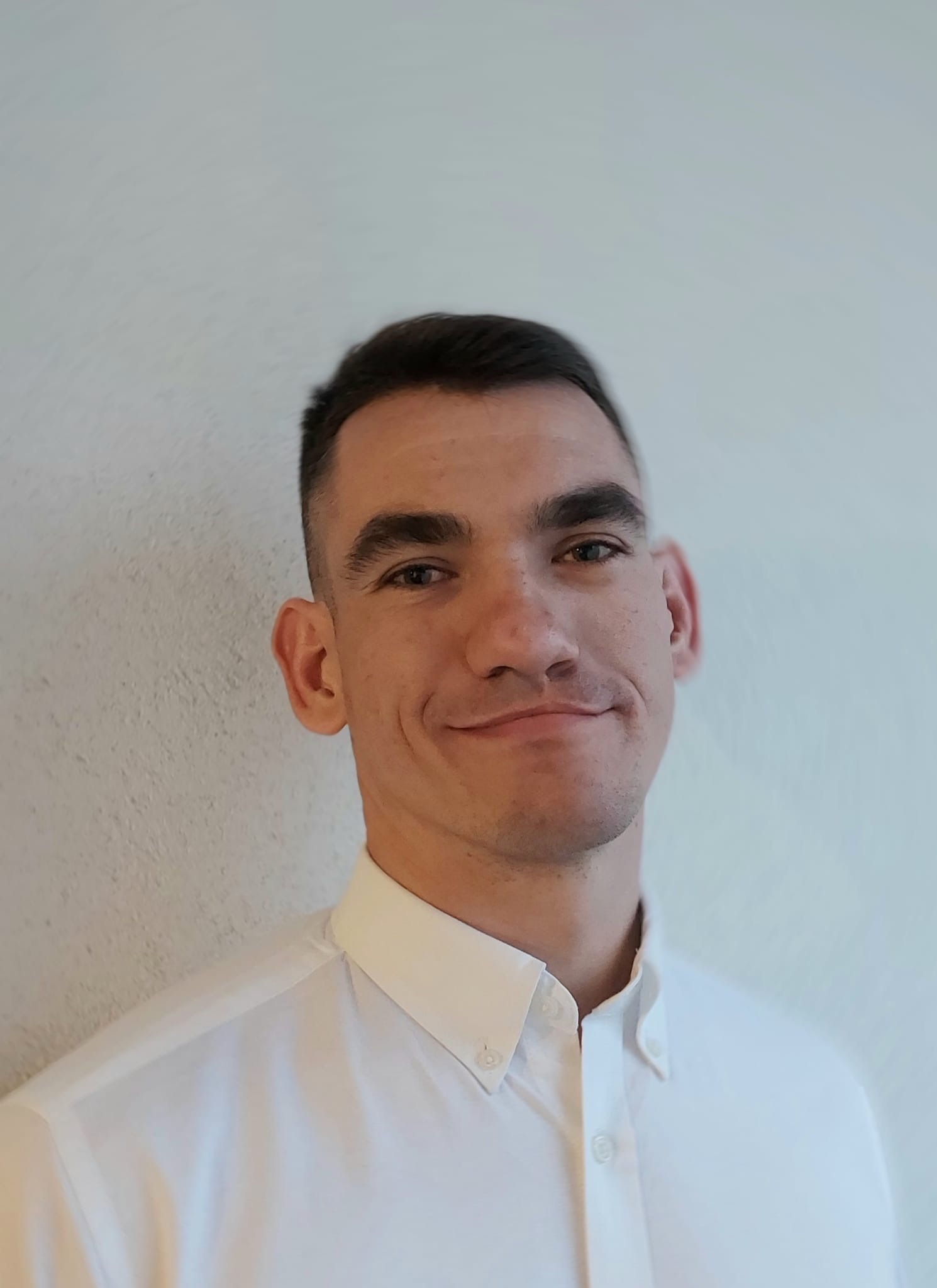 Portret młodego mężczyzny w białej koszuli na tle jasnej ściany. Krótkie, ciemne włosy, lekki uśmiech, wzrok skierowany w obiektyw. Dobre oświetlenie, naturalny wygląd.