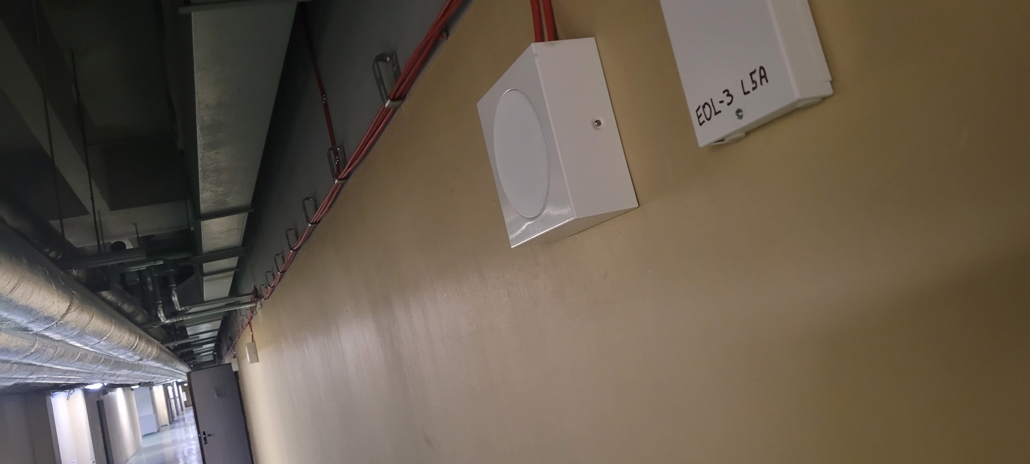 Białe głośniki ewakuacyjne zamontowane na ścianie korytarza, widoczne przewody w uchwytach. Na obudowie głośnika czarny napis 'EOL-3 L5A'.