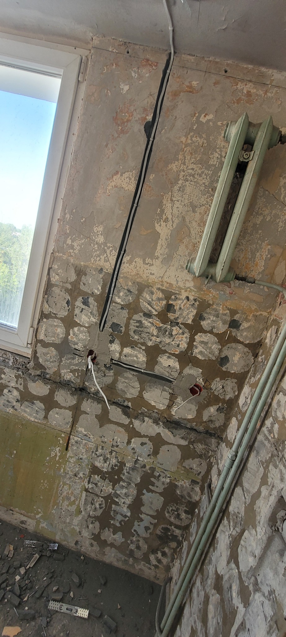 Remont instalacji elektrycznej w Łodzi: widoczne kable w ścianie, grzejnik i okno. Ściana w trakcie renowacji, z odsłoniętą warstwą cegieł.
