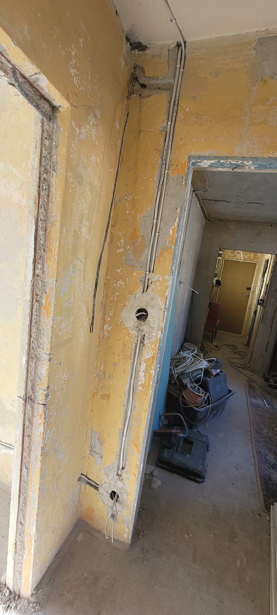 Żółta ściana w trakcie remontu z widocznymi przewodami elektrycznymi, puszkami i narzędziami. Widoczne ślady po farbie i uszkodzenia tynku.