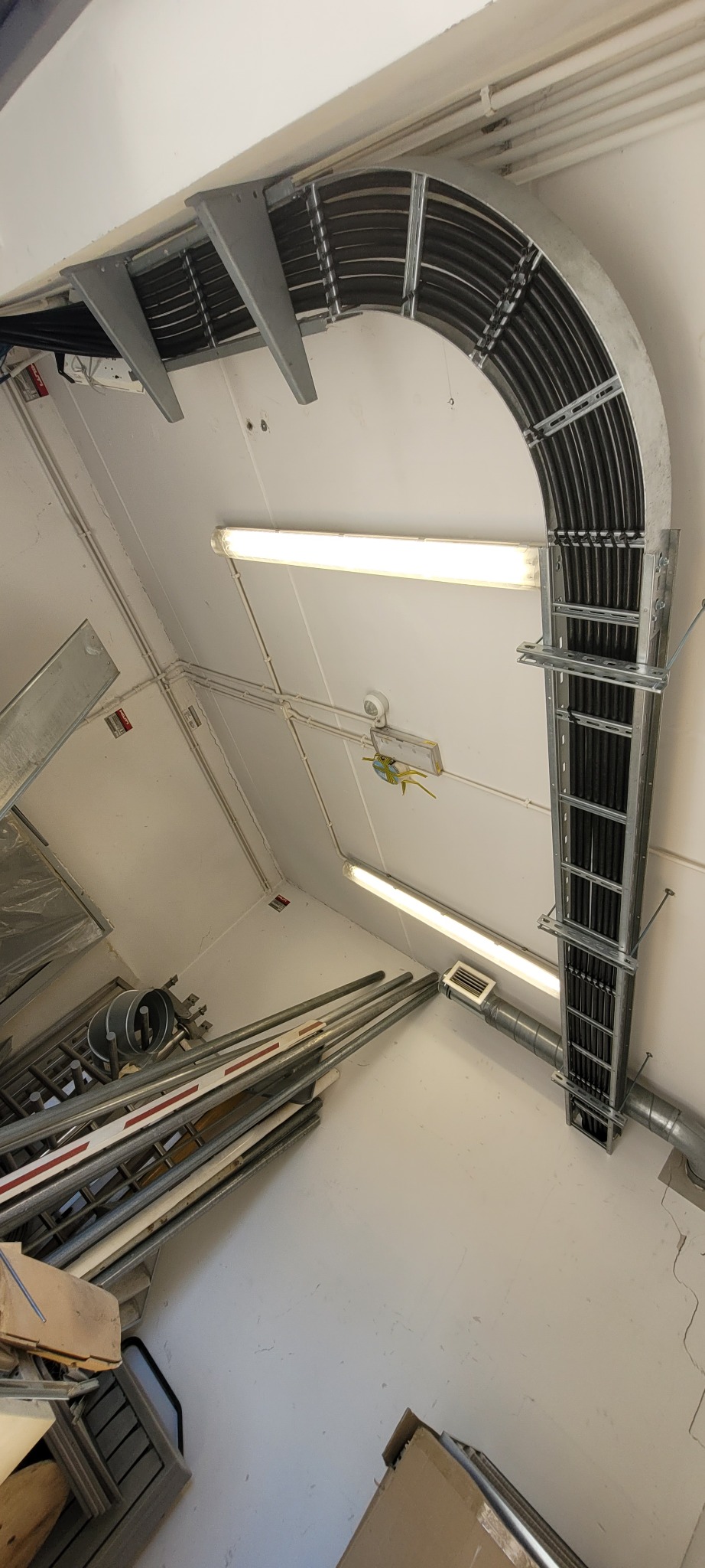 Metalowa trasa kablowa z ułożonymi przewodami, zamontowana pod sufitem. Widoczne elementy mocujące i fragmenty instalacji elektrycznej.