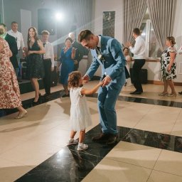 Moje Kadry - Elegancki mężczyzna w błękitnym garniturze tańczy z małą dziewczynką na parkiecie podczas przyjęcia weselnego, w tle bawią się goście.