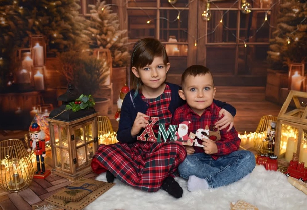Uśmiechnięte rodzeństwo w świątecznej scenerii z drewnianym napisem 'XMAS' na tle bożonarodzeniowej dekoracji i lampionów.