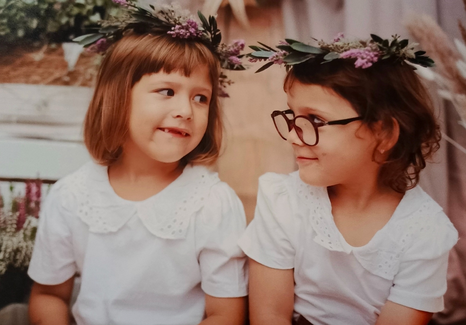 Dwie dziewczynki w białych bluzkach i wiankach z fioletowych kwiatów na głowach, siedzą obok siebie i patrzą na siebie. Jedna ma okulary w brązowych oprawkach.
