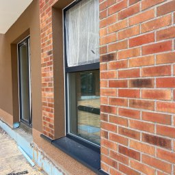 SBS Oleh Liapalo - Fragment elewacji budynku z ceglaną ścianą i oknem zabezpieczonym folią, obok ściana z brązowym tynkiem strukturalnym. Widoczne fragmenty ocieplenia fundamentów.