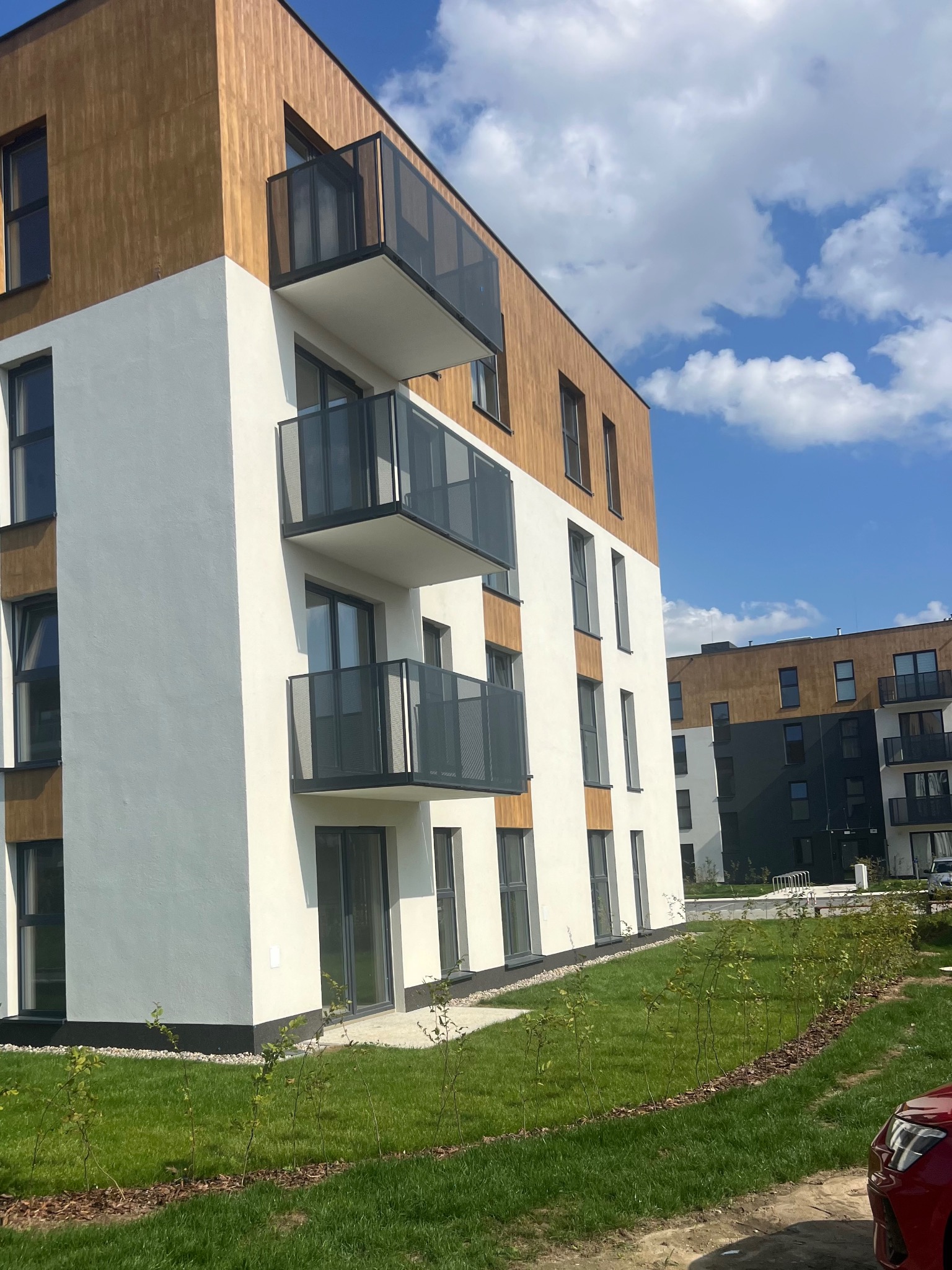 Nowoczesny budynek wielorodzinny z balkonami i elewacją w kolorze białym i drewnopodobnym, na tle błękitnego nieba i zielonego trawnika.