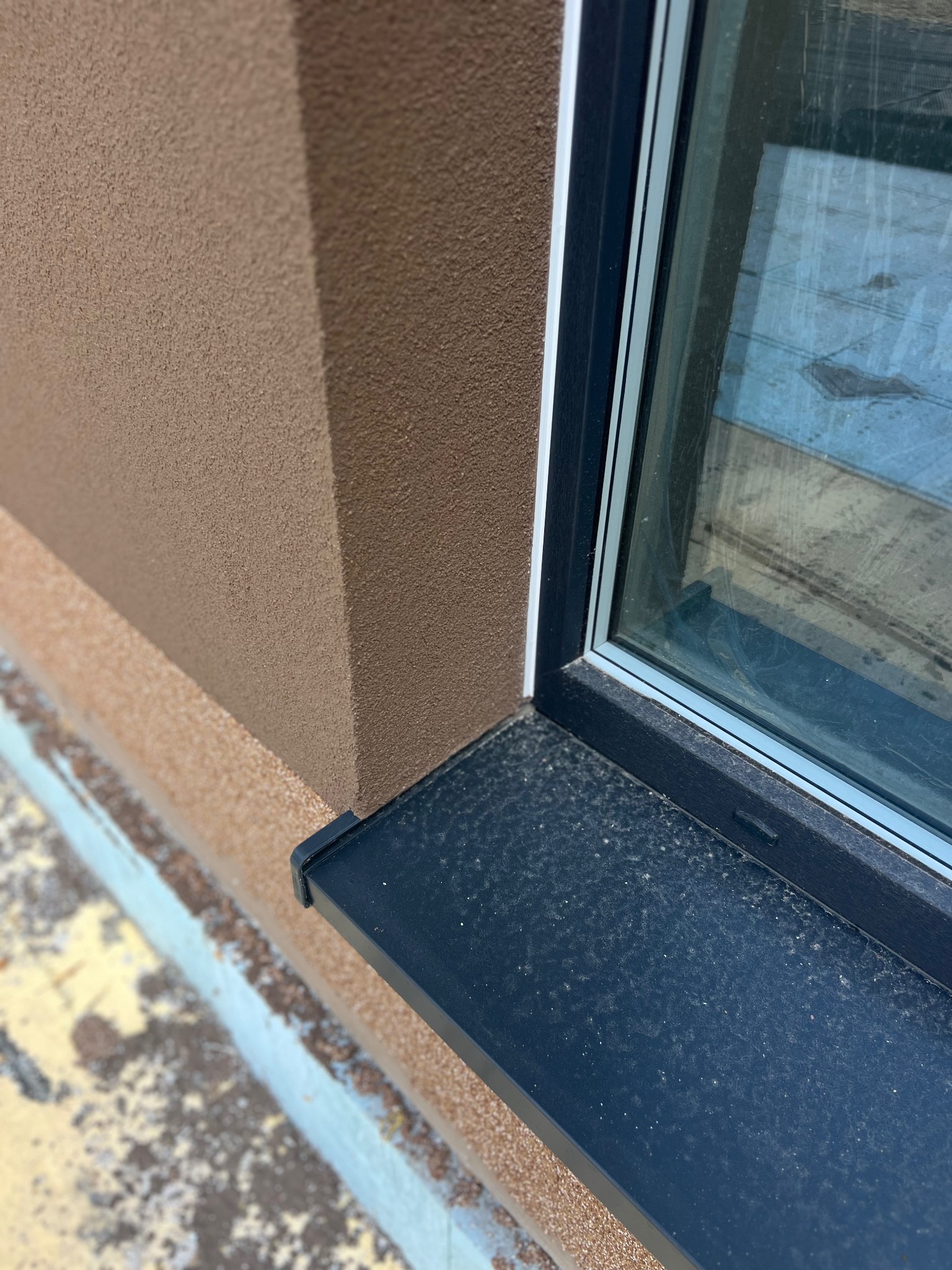 Fragment elewacji z oknem: brązowy tynk strukturalny, czarny parapet. Widoczne zabrudzenia na parapecie i dolnej części elewacji. Ujęcie z dołu, podkreśla fakturę.