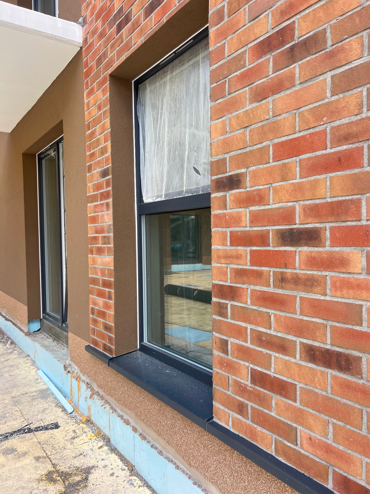 Fragment elewacji budynku z ceglaną ścianą i oknem zabezpieczonym folią, obok ściana z brązowym tynkiem strukturalnym. Widoczne fragmenty ocieplenia fundamentów.