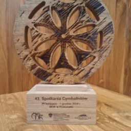 Drewniana statuetka z ażurowym wzorem kwiatowym, na podstawie z napisem '43. Spotkania Cymbalistów', na tle drewnianej ściany. Unikatowy design i naturalne materiały.